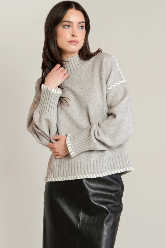 A woman wearing a light gray knit mock-neck sweater with white contrast trim and a black faux-leather skirt, standing against a plain light beige studio background.