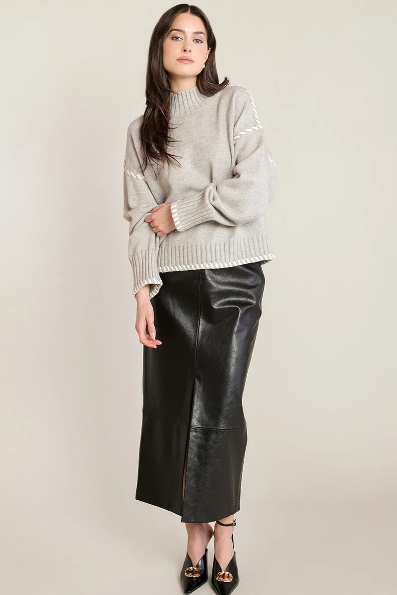 A woman modeling a light gray knit mock-neck sweater with decorative sleeve details and a black leather midi skirt, paired with black pointed-toe heels, against a plain light gray studio background.