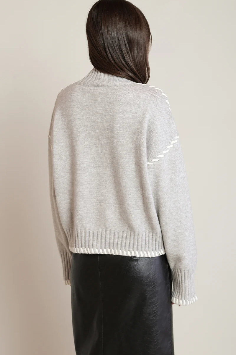 A woman shown from the back wearing a light gray knit sweater with white contrast stitching details and a black leather skirt, set against a plain beige studio background.