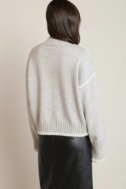 A woman shown from the back wearing a light gray knit sweater with white contrast stitching details and a black leather skirt, set against a plain beige studio background.