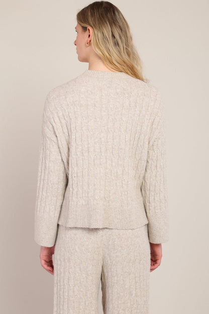 A woman shown from the back wearing a light beige cable-knit sweater and matching knit pants against a plain light beige studio background.