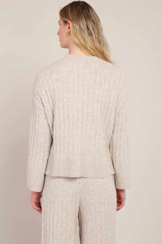 A woman shown from the back wearing a light beige cable-knit sweater and matching knit pants against a plain light beige studio background.
