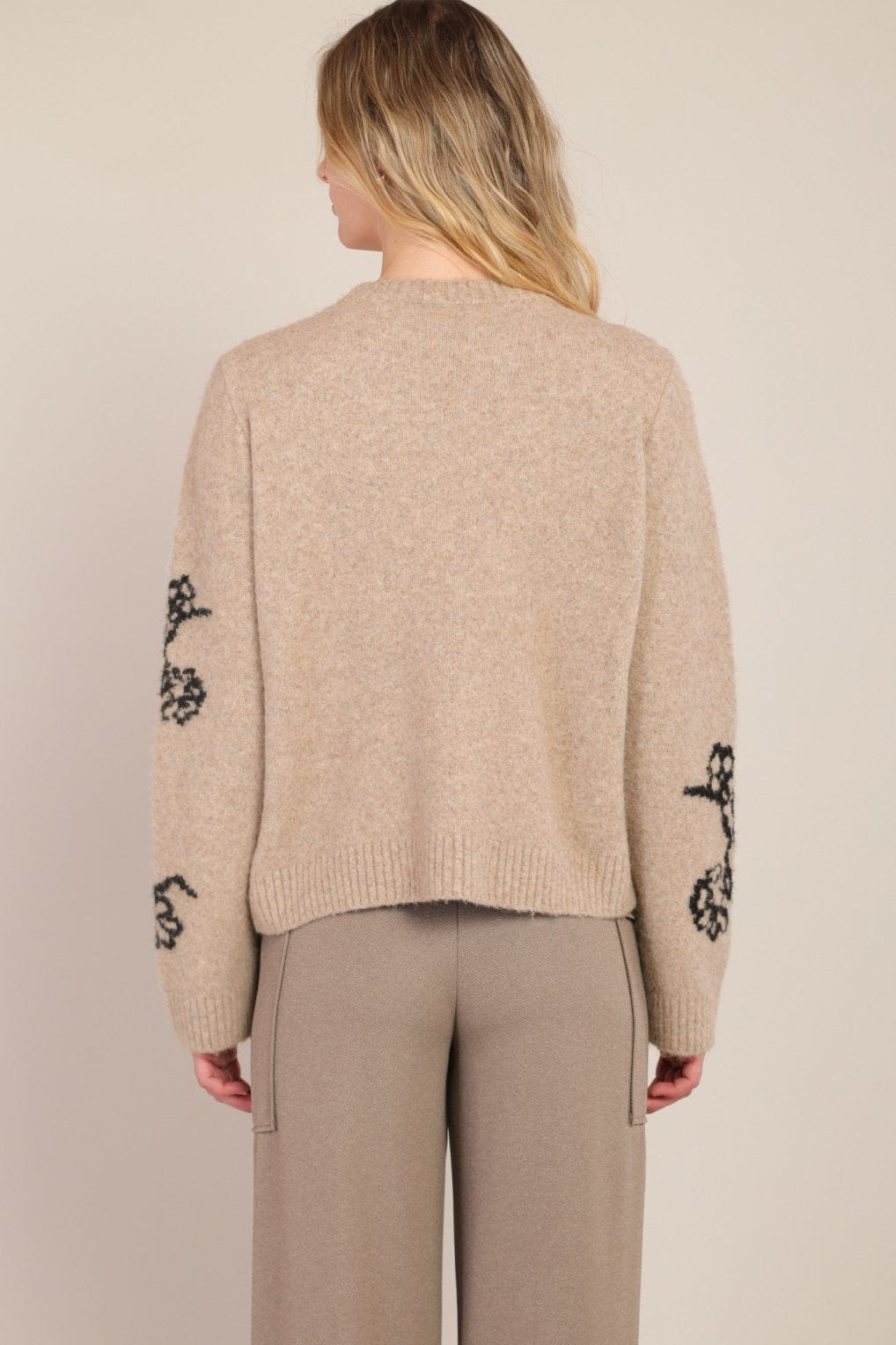 Beige sweater with black floral patterns on sleeves worn by a person against a beige background
