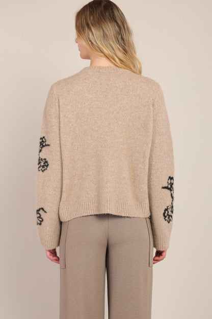 Beige sweater with black floral patterns on sleeves worn by a person against a beige background