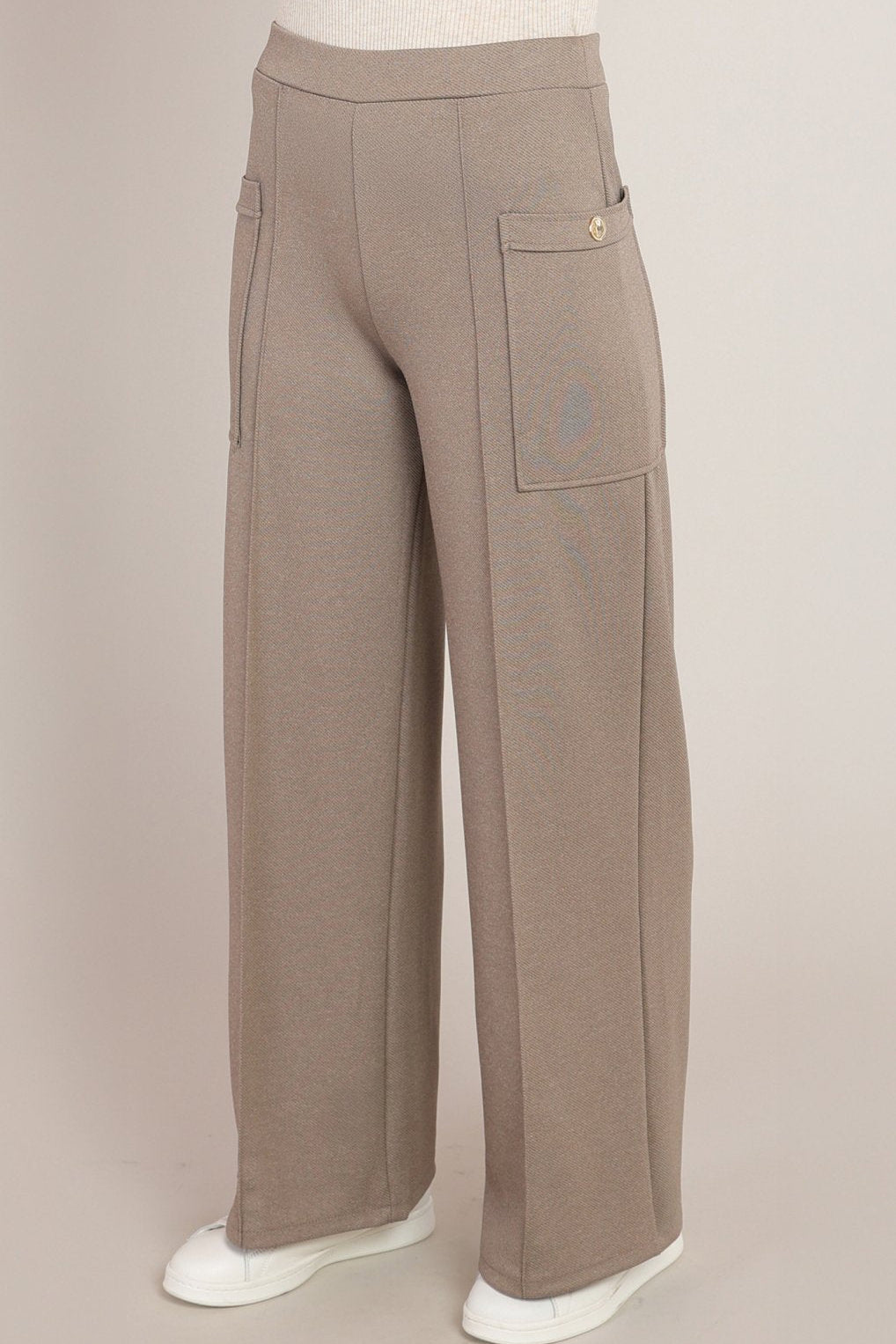 A pair of wide-leg taupe pants displayed on a mannequin lower body, featuring a high waist, side cargo pocket with a gold button, and white sneakers against a solid light beige background.