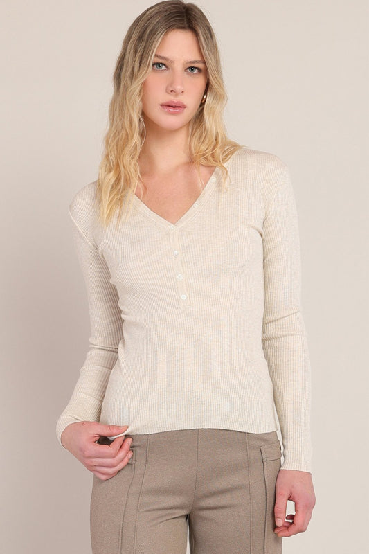 A woman modeling a cream-colored ribbed knit long-sleeve V-neck top with a three-button placket, paired with taupe pants, against a solid light beige background.