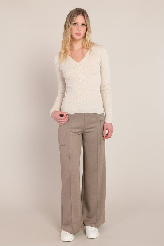 A woman modeling a cream long-sleeve knit top and taupe wide-leg trousers against a plain beige studio background.