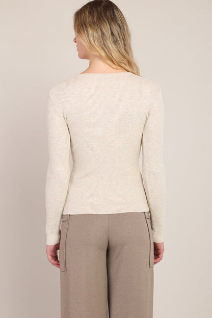 A woman shown from the back wearing a fitted long-sleeve knit sweater in a light cream color and taupe trousers, set against a plain light beige studio background.
