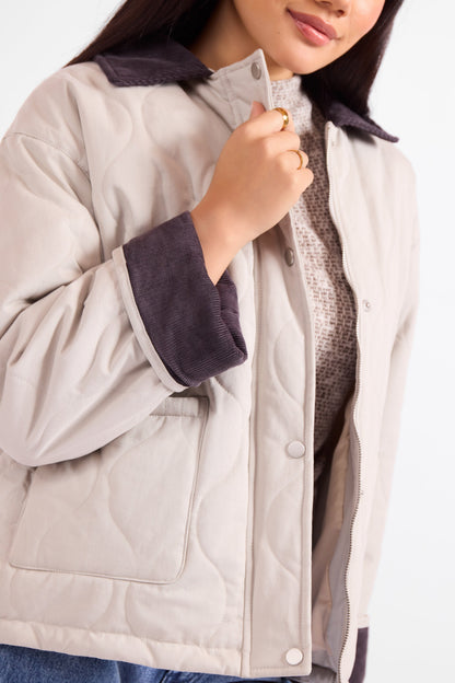 A close-up of a woman wearing a light beige quilted jacket with dark brown corduroy collar and cuffs, layered over a textured knit sweater.