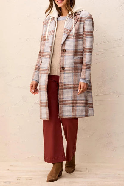 A woman models a mid-length hooded plaid coat in beige, brown, and white, worn over a beige top and rust-colored wide-leg pants, against a textured beige wall.