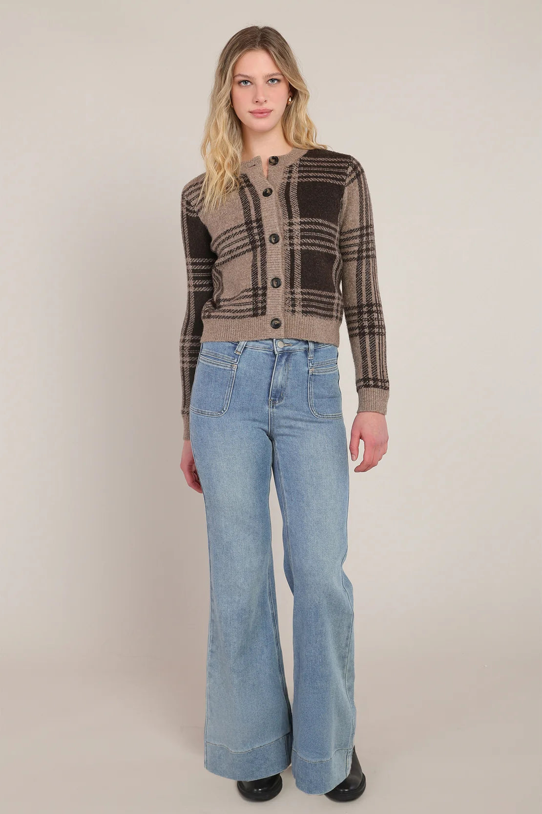A woman modeling a brown and beige plaid button-up cardigan sweater paired with light-wash blue flare jeans, standing against a neutral beige studio background.