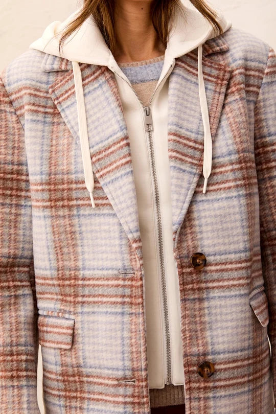 A close-up of a person wearing a hooded zip-front jacket with a soft plaid pattern in beige, blush pink, and muted blue, layered over a cream zip hoodie.
