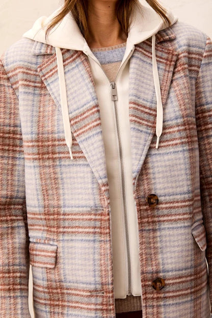 A close-up of a person wearing a hooded zip-front jacket with a soft plaid pattern in beige, blush pink, and muted blue, layered over a cream zip hoodie.