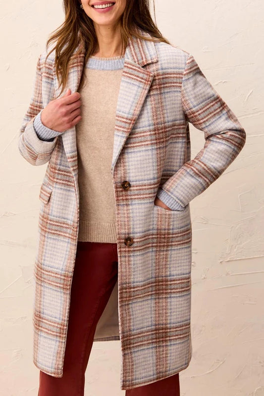 A woman wearing a long plaid coat over a beige sweater and rust red pants, standing against a textured beige wall.