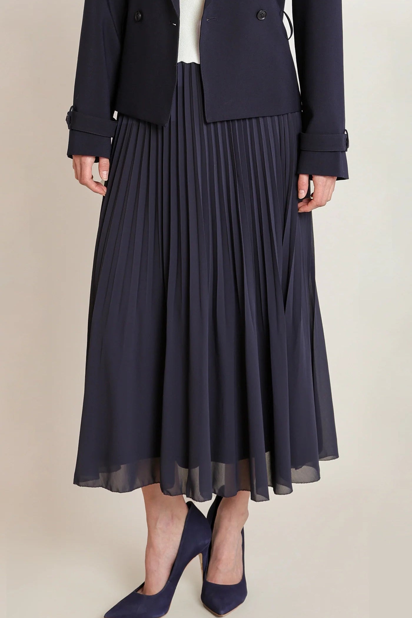 A model wearing a dark navy blue pleated midi skirt, paired with a matching navy blazer and navy blue pointed-toe heels, set against a plain beige studio background.