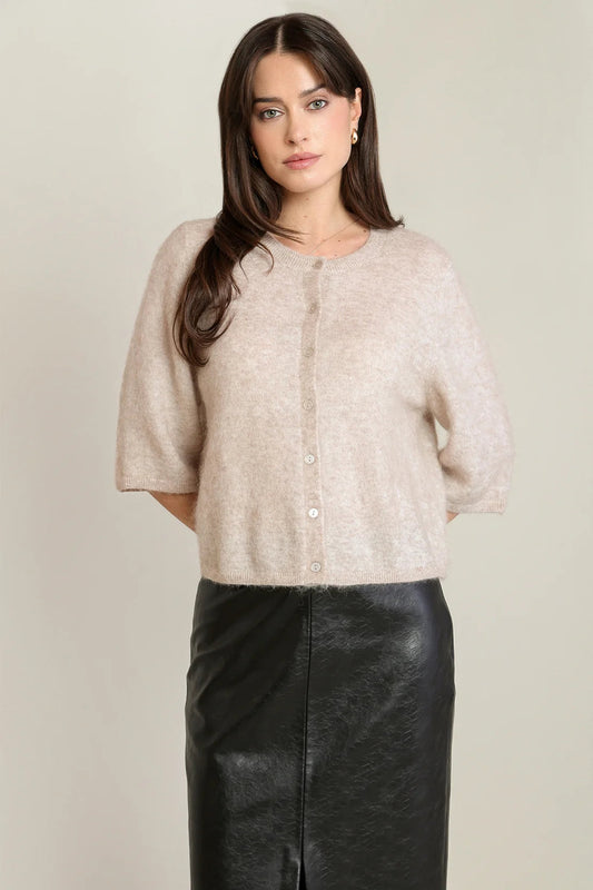 A woman modeling a light beige short-sleeve knit cardigan with a button-up front, paired with a black leather skirt, against a plain light gray studio background.