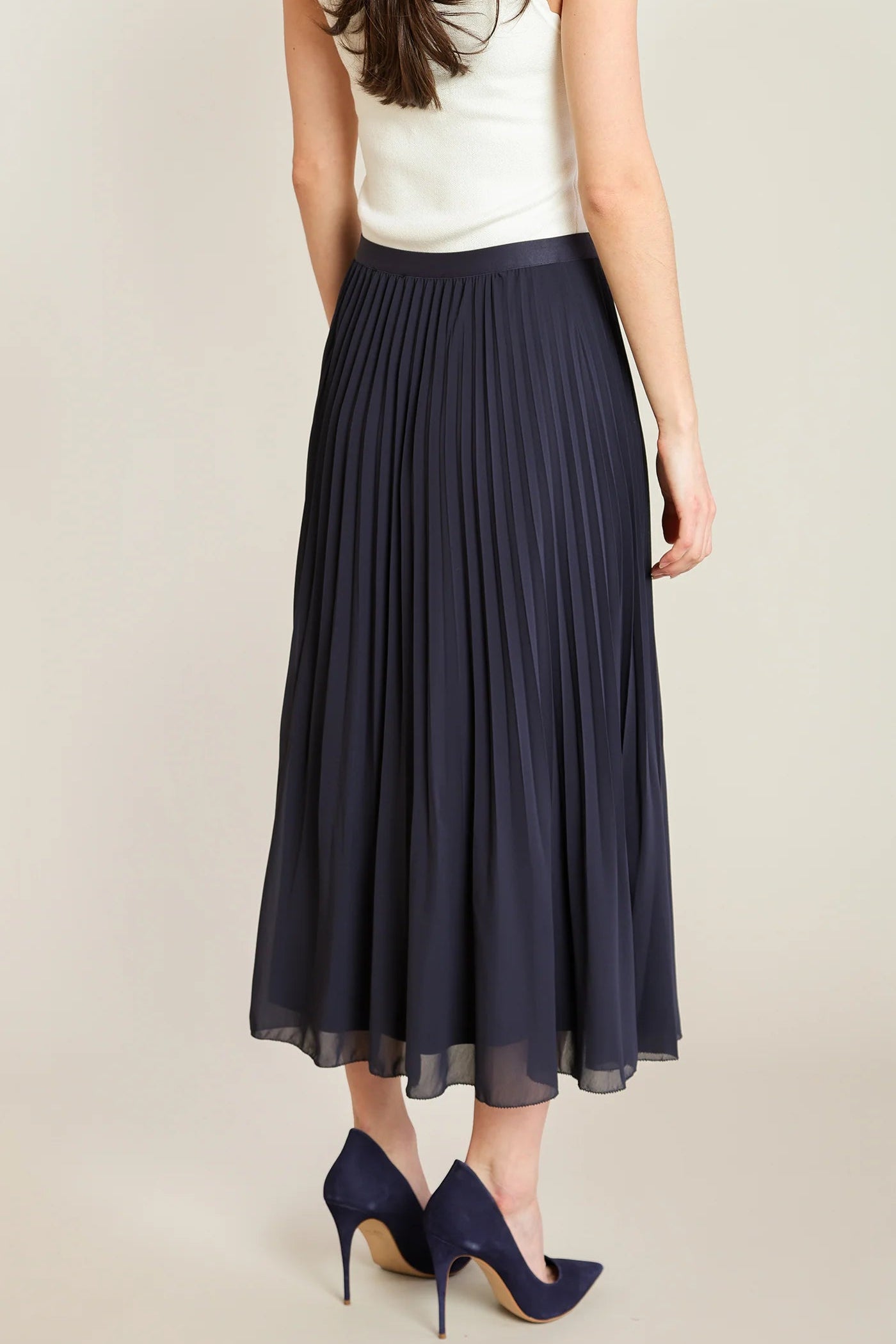 A woman wearing a navy blue pleated midi skirt, a white sleeveless top, and navy blue high heels, shown from the back against a light beige studio background.