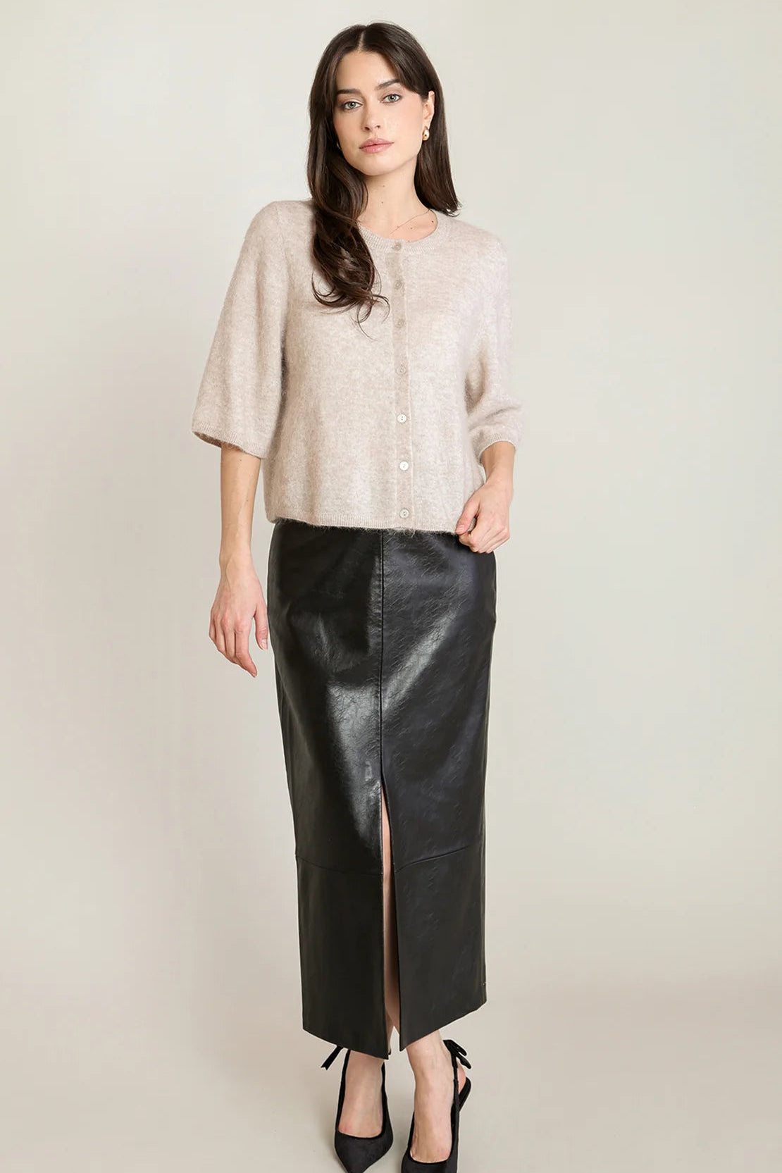 A woman modeling a light beige textured short-sleeve button-up top paired with a black faux-leather midi skirt featuring a front slit, set against a plain light beige studio background.