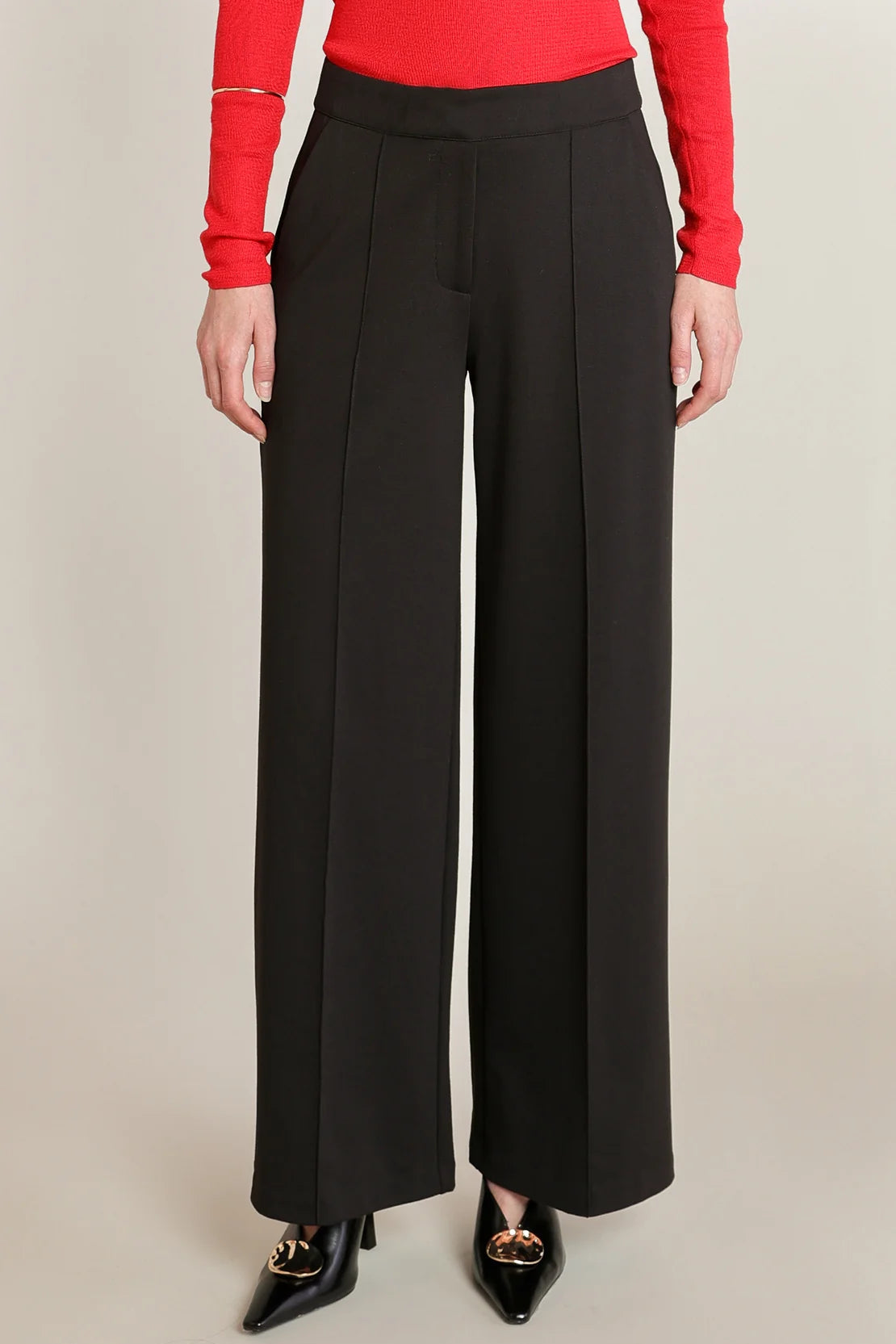 A model wearing black wide-leg trousers with a red long-sleeve top and black pointed-toe heels, shown from the waist down against a neutral beige background.