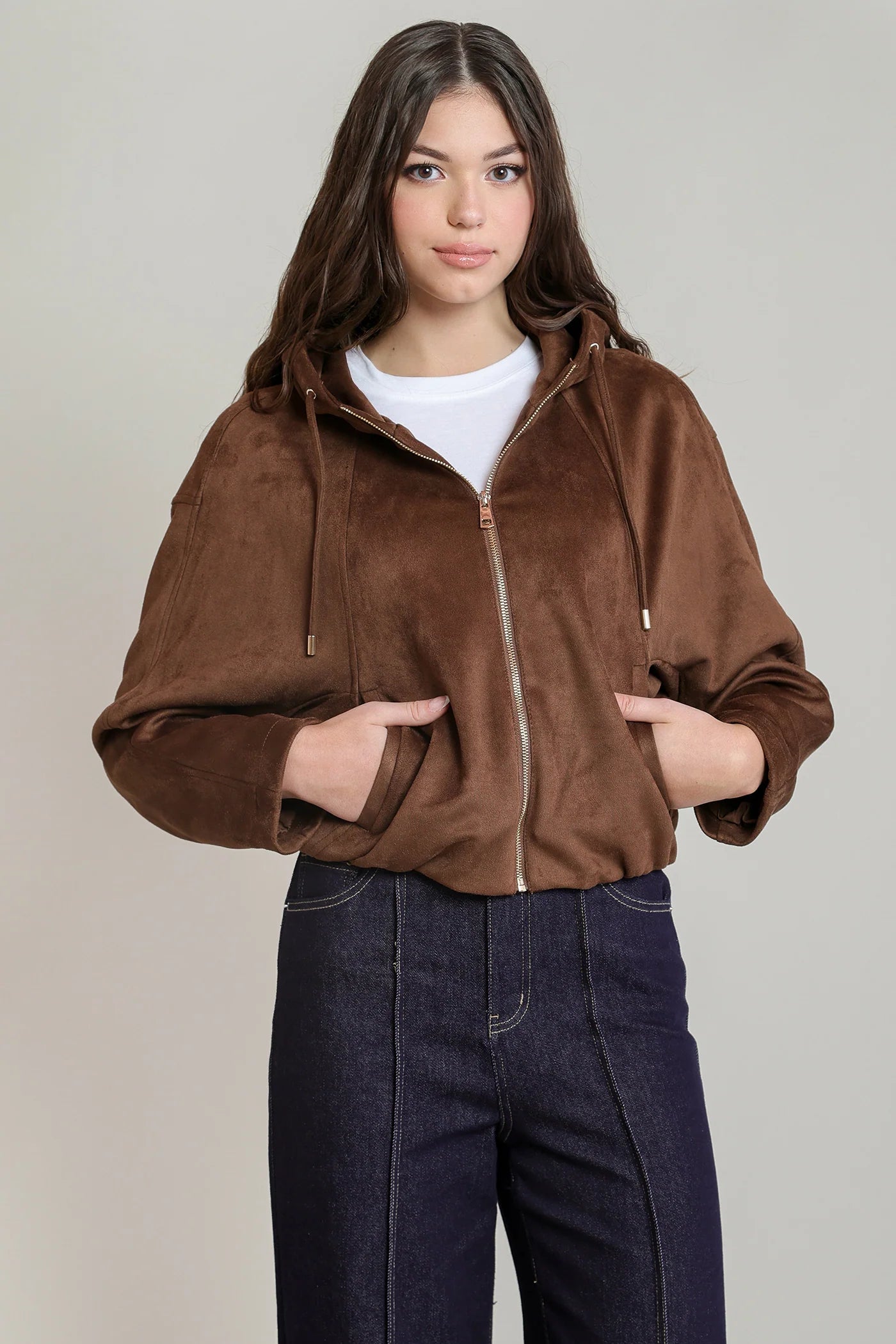 A woman modeling a cropped brown faux-suede zip-up hoodie jacket with drawstrings and front pockets, paired with a white t-shirt and dark blue jeans, against a solid light gray background.