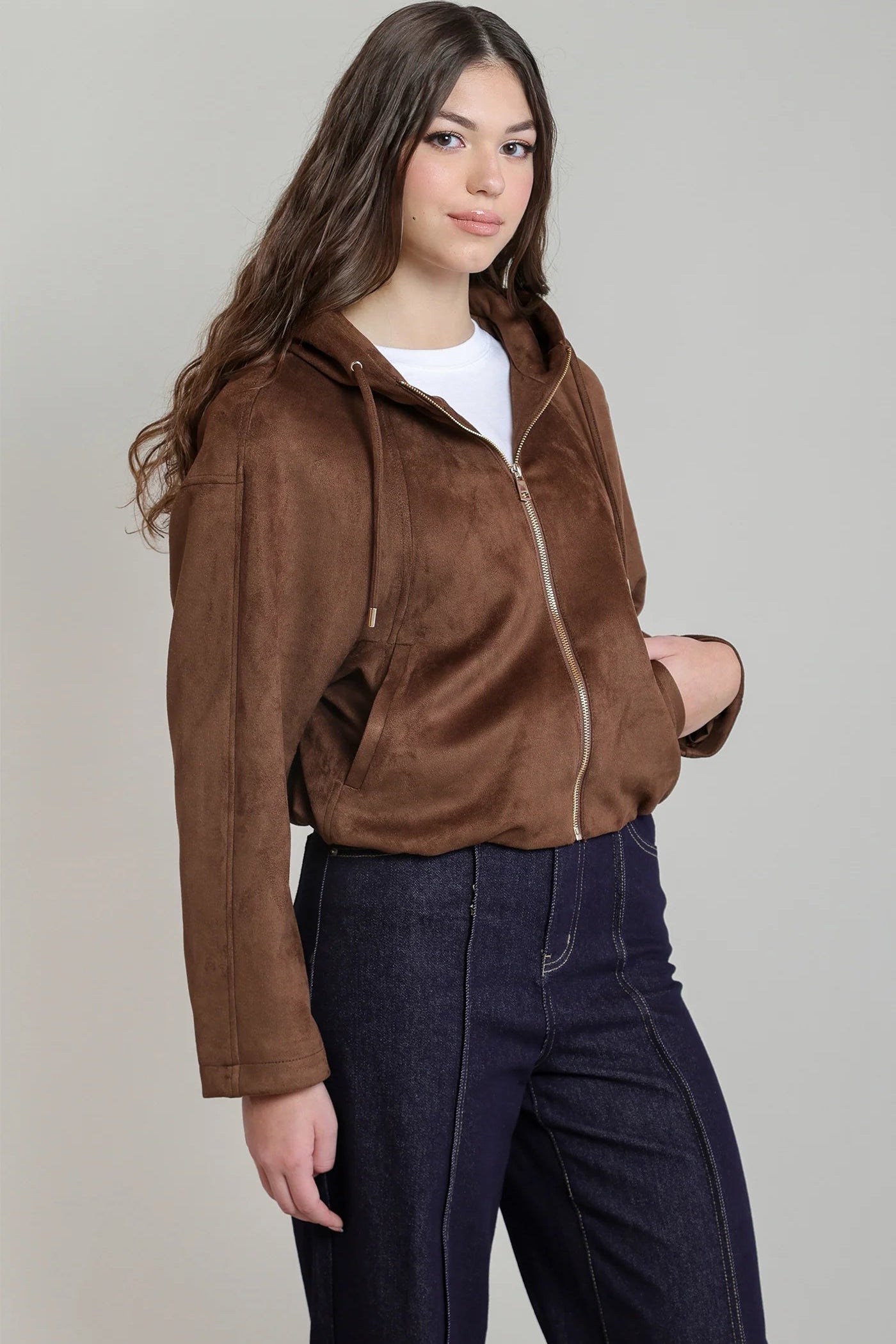 A woman modeling a brown suede-like hooded zip-up jacket over a white t-shirt and dark blue jeans, photographed against a solid light gray studio background.