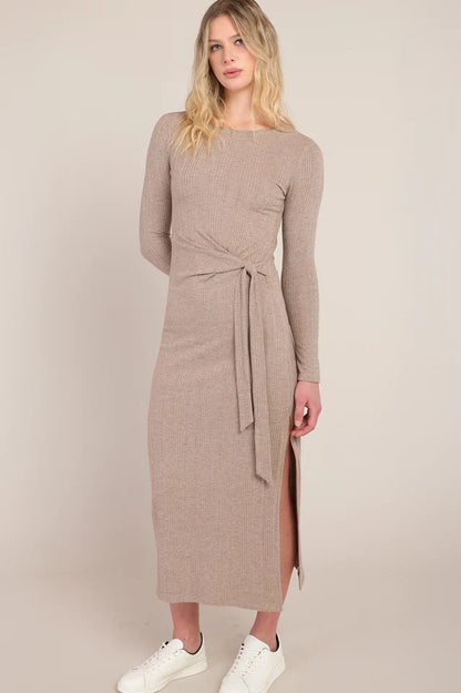 A woman modeling a long-sleeve taupe knit midi dress with a tie waist and side slit, paired with white sneakers, against a solid beige studio background.