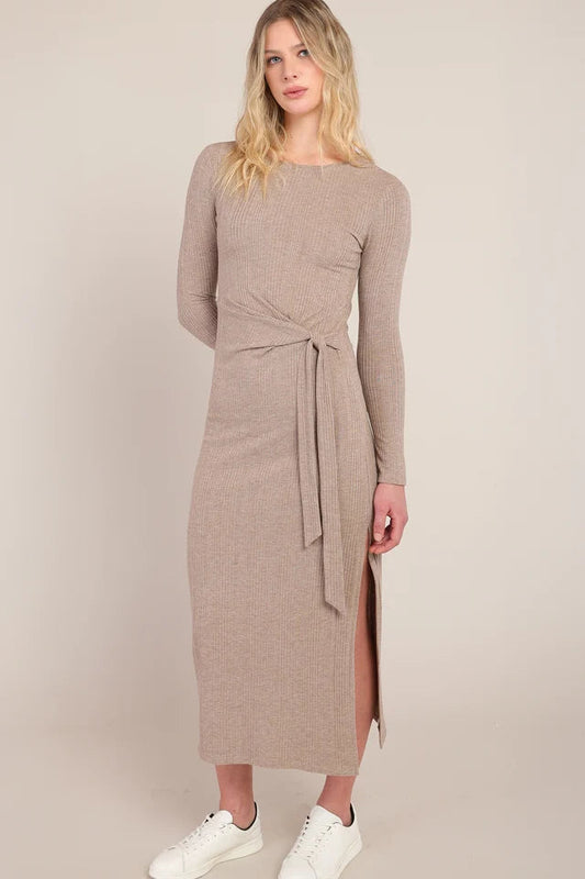 A woman modeling a long-sleeve taupe knit midi dress with a tie waist and side slit, paired with white sneakers, against a solid beige studio background.