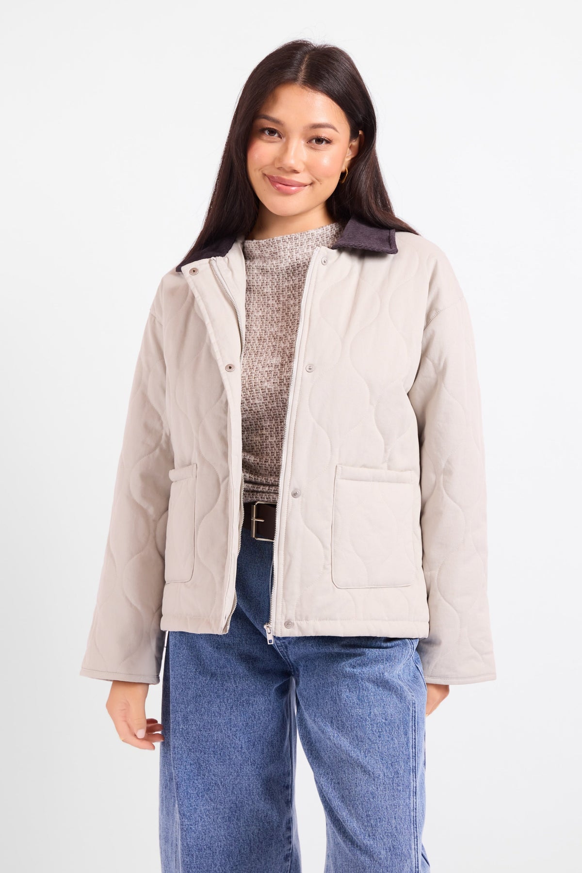 A woman modeling a light beige quilted jacket with a dark brown collar, worn over a textured knit top and blue jeans, against a plain white studio background.