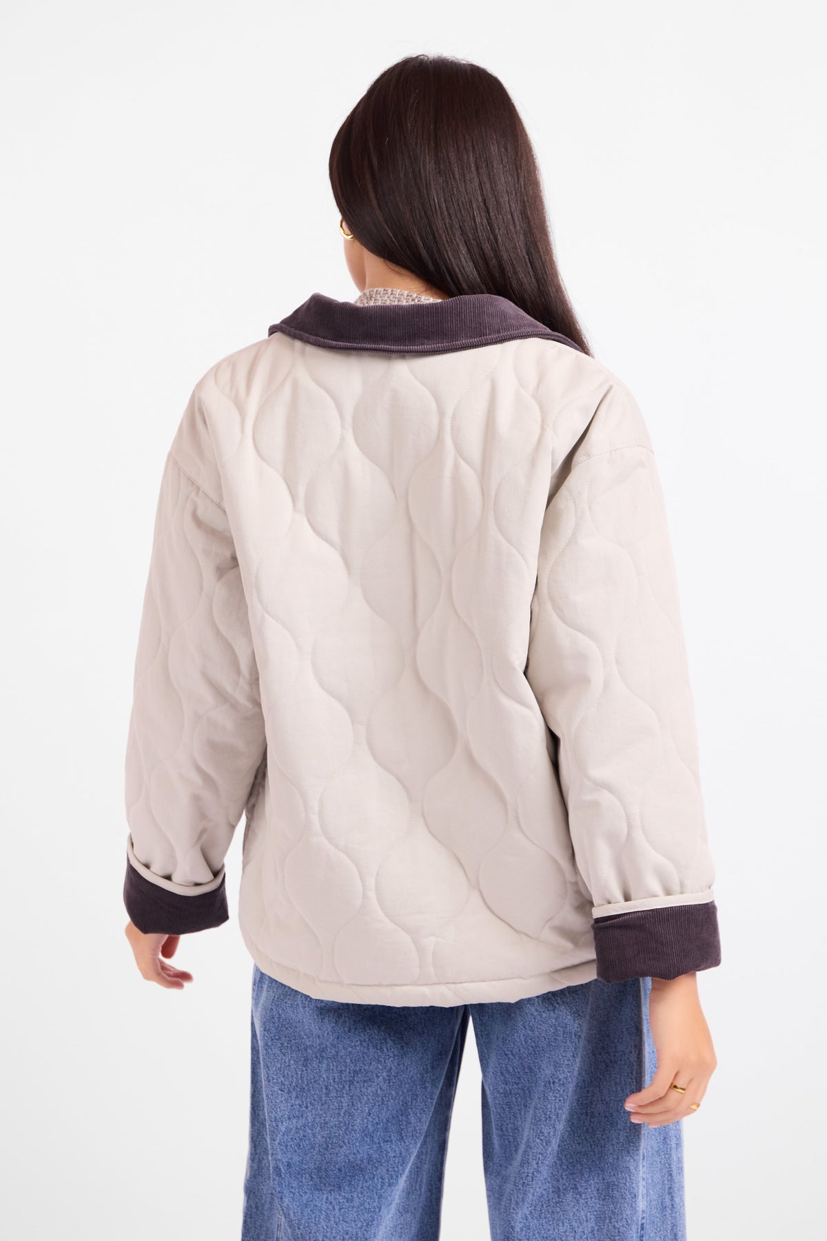 A woman shown from the back wearing a quilted jacket with a light beige body and dark brown trim on the collar and cuffs, paired with blue jeans against a plain white background.