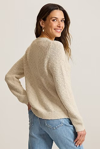A woman wearing a cream-colored, textured knit sweater and blue jeans, photographed from a three-quarters back angle against a light gray studio background.