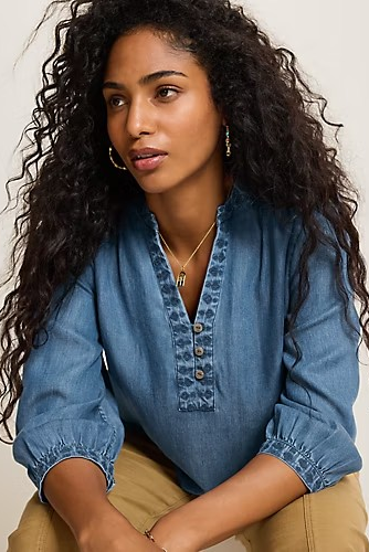 A woman with long, curly dark hair wearing a blue denim-style blouse with a buttoned split neckline and gathered sleeves, paired with tan pants, posed against a plain light background.
