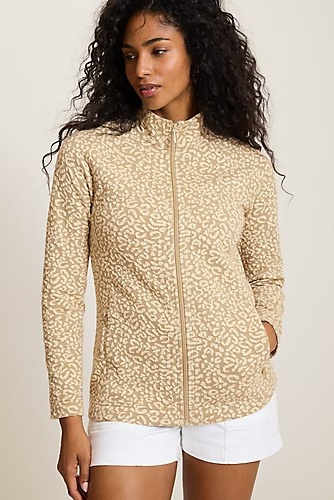 A woman wearing a beige and tan leopard-print zip-up jacket with white shorts, standing against a plain light beige studio background.